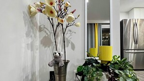 A room with a tall vase containing flowers and a teddy bear, a potted plant, and a kitchen area with a refrigerator.