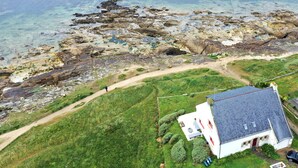 A coastal house with a paved path leading to the sea, surrounded by greenery and a clear view of the rocky shoreline.