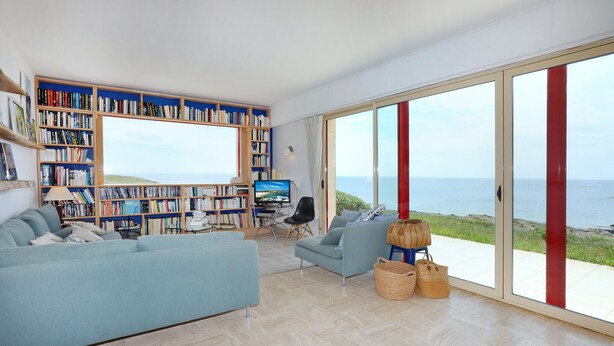 A spacious living room with a large bookshelf, a television, and a view of the sea through large windows.