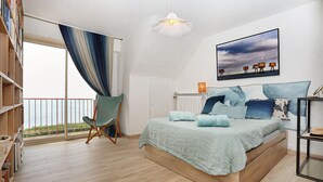 A bedroom with a large bed, a chair, a bookshelf, a radiator, and a view of the sea.
