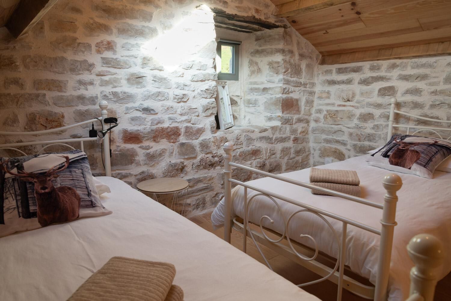 A rustic stone-walled room with two beds, each with a wooden headboard and a pillow with a deer print.