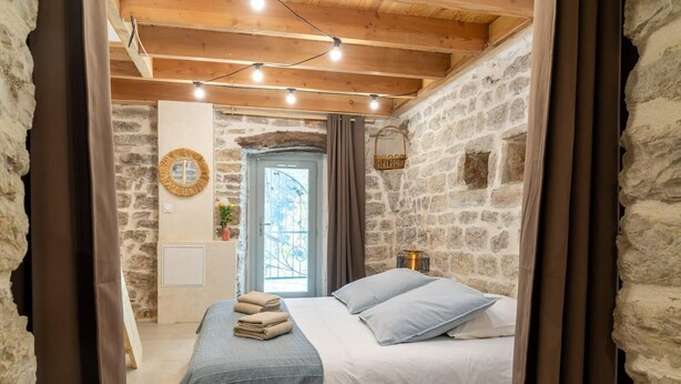 A cozy bedroom with a stone wall, a bed with a blue blanket, a wooden ceiling, and a small window.