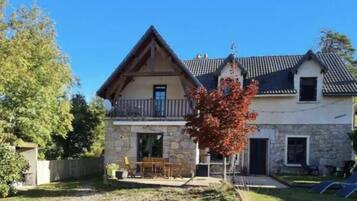 Une maison en pierre à deux étages, dotée d’un balcon, d’une petite remise attenante et d’une pelouse aménagée avec du mobilier d’extérieur.