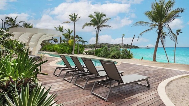Un pontile di legno con poltrone relax e una piscina, circondato da palme e vista sull'oceano.