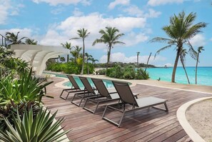 Une terrasse en bois avec des chaises longues et une piscine, entourée de palmiers et offrant une vue sur l’océan.