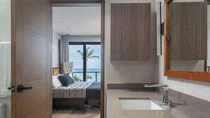 Una camera d'albergo moderna con un letto, una grande finestra vista mare, bagno con lavandino e specchio, e mobili in legno.