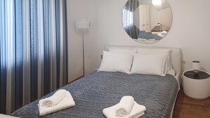 Ein ordentlich bezogenes Bett mit einer grauen, gemusterten Tagesdecke und zwei sauber gefalteten weißen Handtüchern. Ein runder Spiegel hängt über dem Bett und spiegelt einen Teil des Raumes wider.