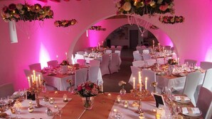 Une salle de banquet avec des tables rondes dressées pour un événement formel, décorée de compositions florales et de bougies.