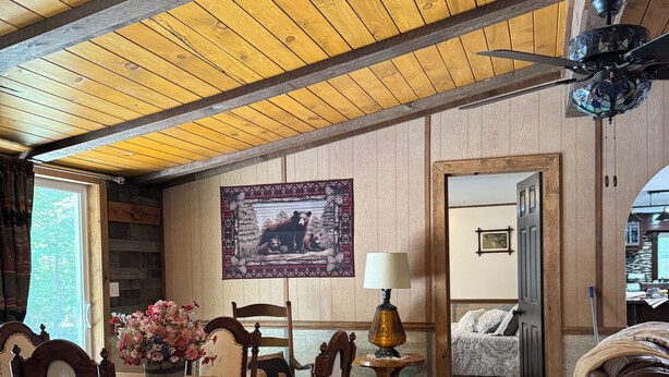 Una habitación acogedora con vigas de madera en el techo, una zona de comedor con mesa y sillas de madera, una cama con cabecera y un tapiz decorativo en la pared.