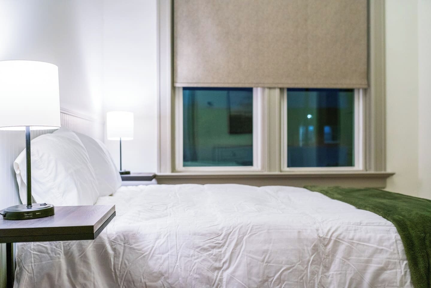 Una camera d'albergo con un letto, un comodino con una lampada, una finestra vista e una coperta verde sul letto.