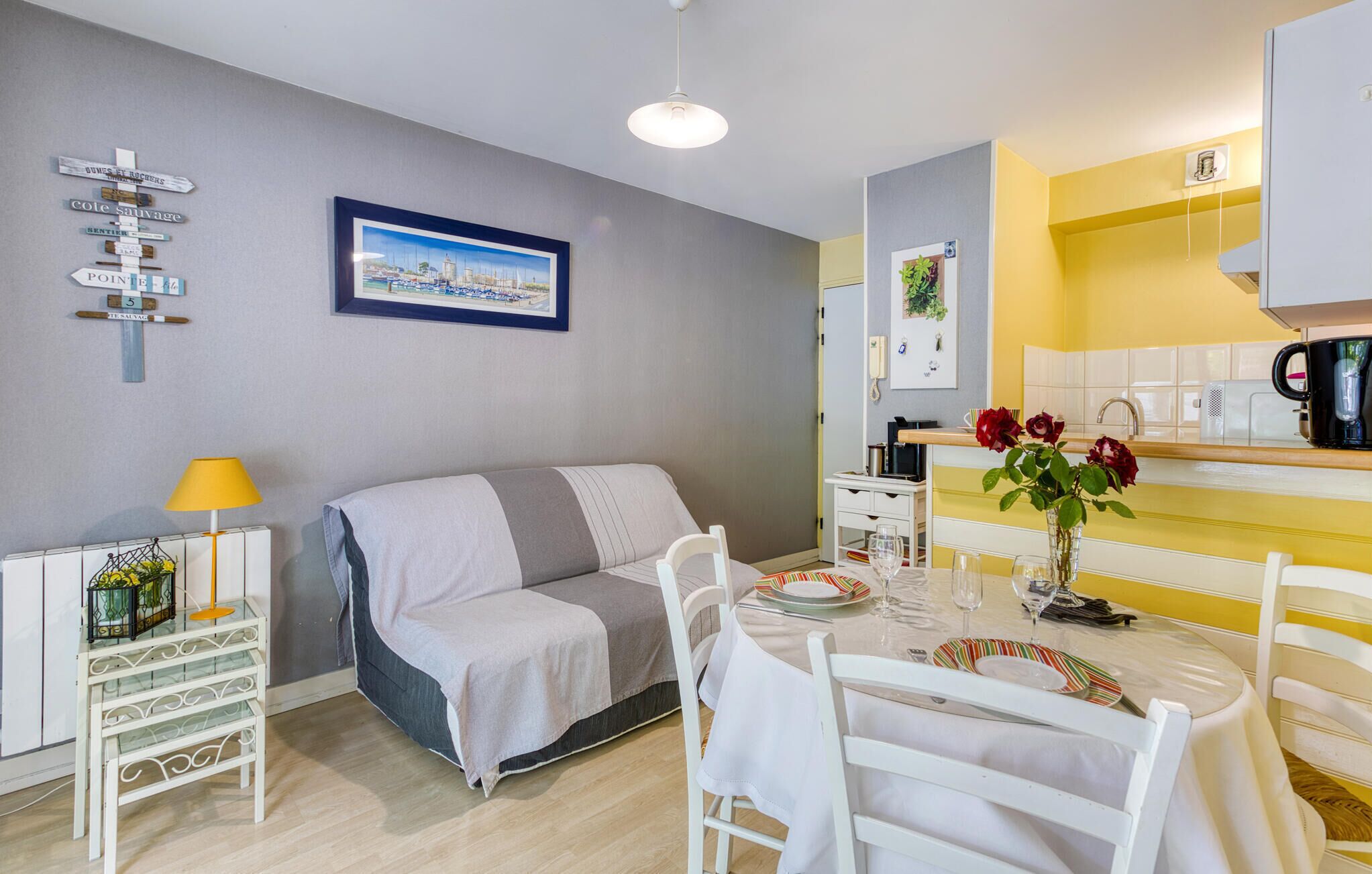 Une pièce confortable comprenant un lit, une table à manger dressée pour deux personnes, un coin cuisine et un mur orné d’un tableau et de panneaux d’indication.