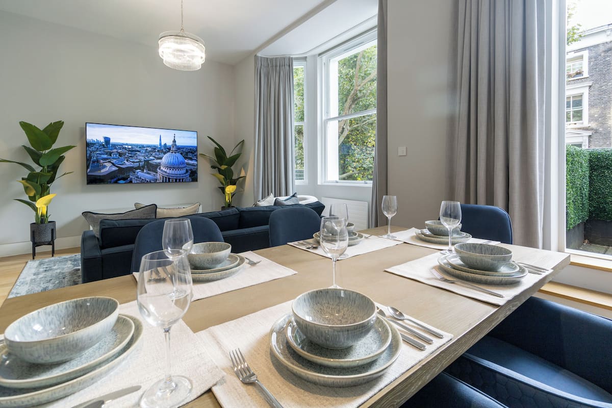 A dining area with a long table set for six, a modern sofa, and a large window with curtains.