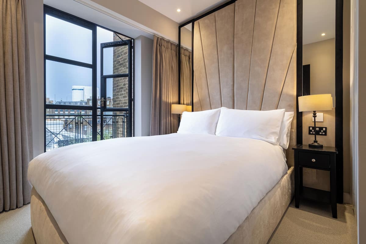 A large bed with white linens, a nightstand with a lamp, and a view of the city through a large window.