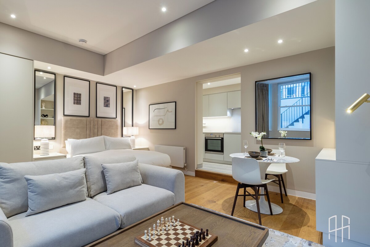 A modern living room with a sofa, a coffee table with a chessboard, a dining area with a table and chairs, and a kitchen in the background.