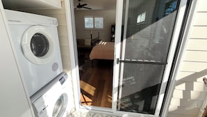 A laundry area with a washer and dryer, a sliding glass door leading to a bedroom with a bed and a ceiling fan. - Spacious Byron Central Studio Apartment w/ Private Balcony (Byron Bay)