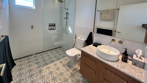 A bathroom with a glass-enclosed shower, a toilet, a sink with a large mirror, patterned floor tiles, and a towel rack. - Spacious Byron Central Studio Apartment w/ Private Balcony (Byron Bay)