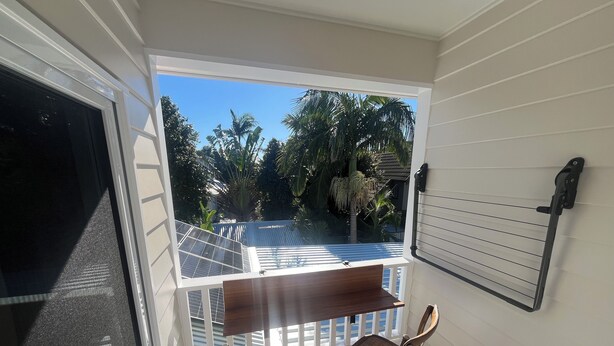 A balcony with a table and two chairs, overlooking a garden with palm trees and a metal roof structure. - Spacious Byron Central Studio Apartment w/ Private Balcony (Byron Bay)