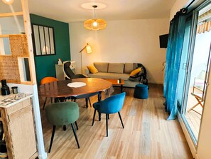 Una sala de estar acogedora con una mesa de comedor de madera, sillas azules, un sofá con cojines amarillos y un otoman azul. Una separador de mimbre divide el comedor del espacio de la sala.