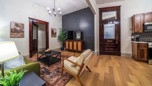 Een woonkamer met een bank, salontafel en een televisie. Er is een keukenhoek met kasten en een magnetron.