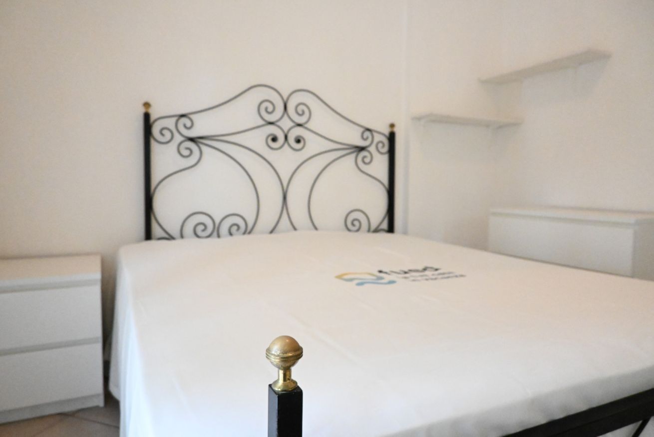 A bedroom with a metal headboard, a bed with a white mattress and a logo, a bedside table, and a white dresser.