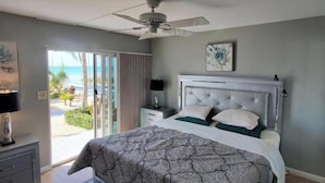 Quarto com cama, ventoinha no teto, mesa de cabeceira, luminária, pintura e vista para a praia.