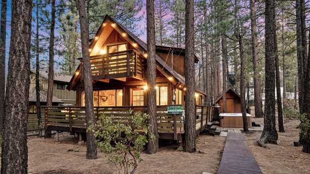 A cabin with a wooden exterior, surrounded by tall pine trees, and a wooden deck leading to the entrance.