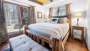A bedroom with a large bed, wooden headboard, two bedside tables with lamps, a bench, and a sliding glass door leading to a balcony.