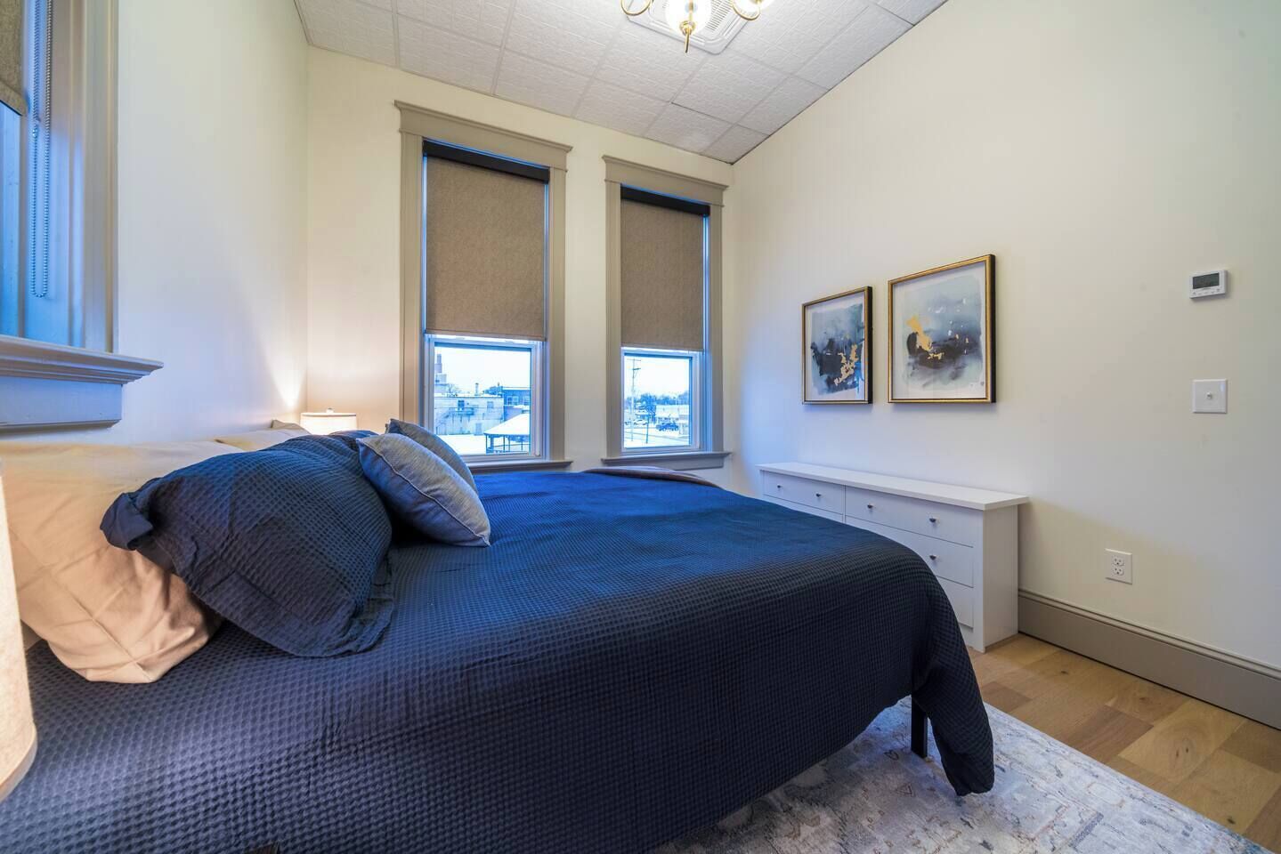 A bedroom with a large bed, two windows, a white dresser, and two framed artworks on the wall.