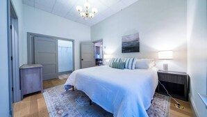 Une chambre avec un grand lit, une table de chevet, une lampe, un tableau accroché au mur et une porte donnant sur une autre pièce.