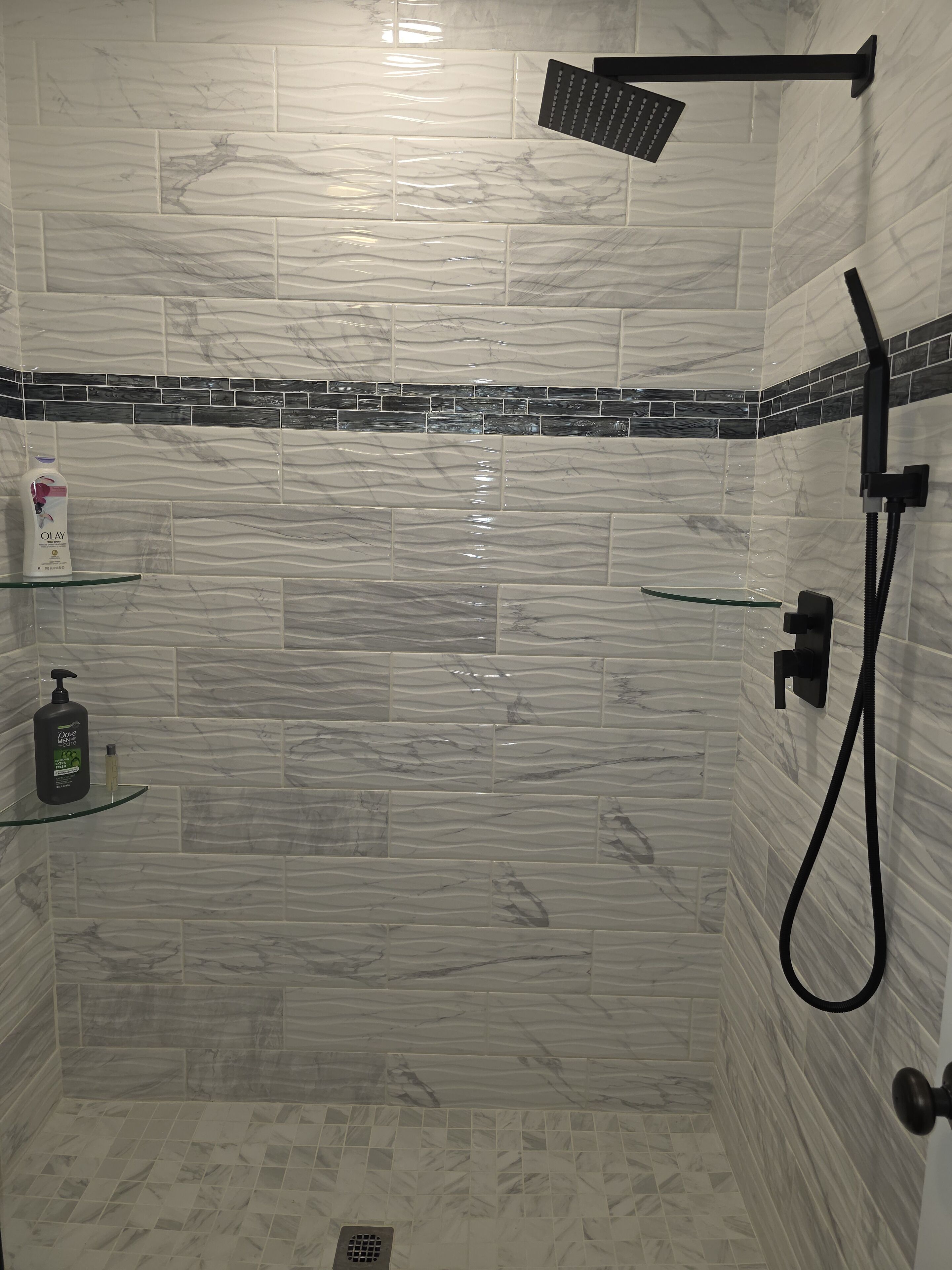 Ducha con cabezal de ducha de mano y grifo empotrado. Dos repisas de vidrio con artículos de aseo.