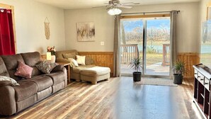 A living room with a brown leather couch, a beige sofa, and a wooden coffee table. There is a sliding glass door leading to an outdoor area with a view of trees and a body of water. - Waterfront 4BR House w/ Sunset View & Fenced Yard (Saint Joseph)