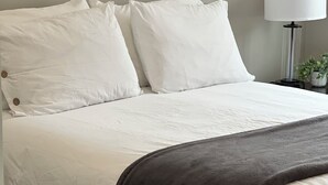 Una cama con ropa de cama blanca y una manta oscura.