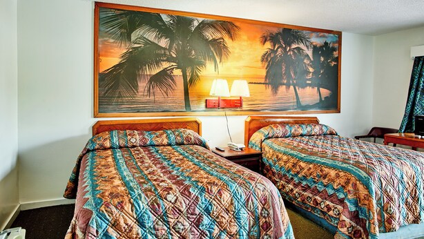 A hotel room with two beds, a painting of a sunset over palm trees, a nightstand with a lamp, and a desk with a chair. - Hospitality Inn (Selma)