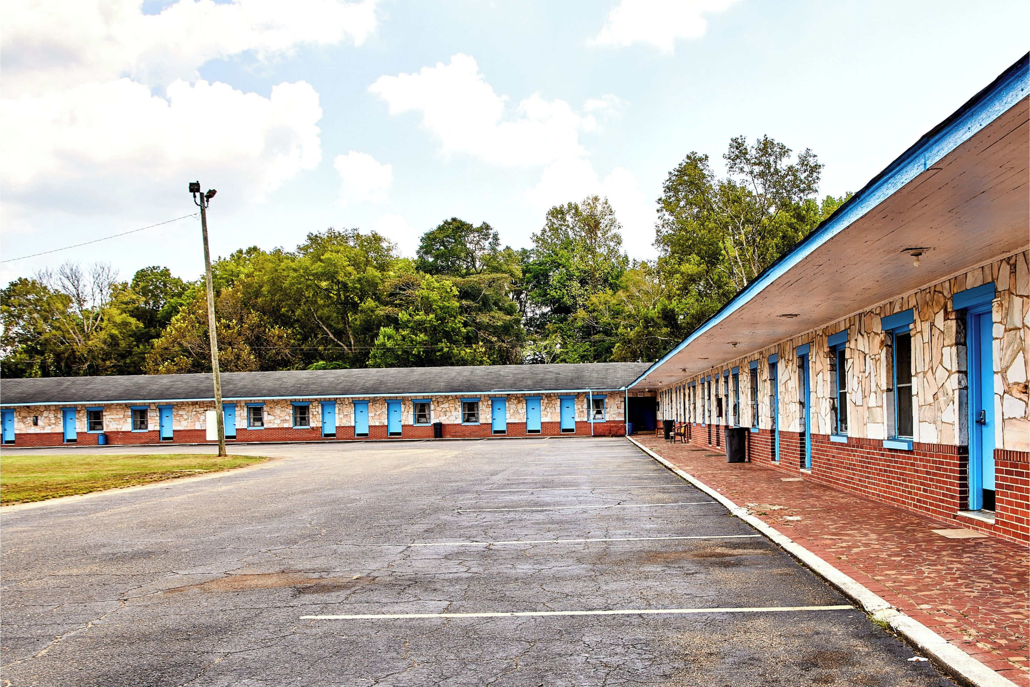 Fila de habitaciones de motel con puertas azules y fachada de ladrillo, un estacionamiento al frente y árboles al fondo.