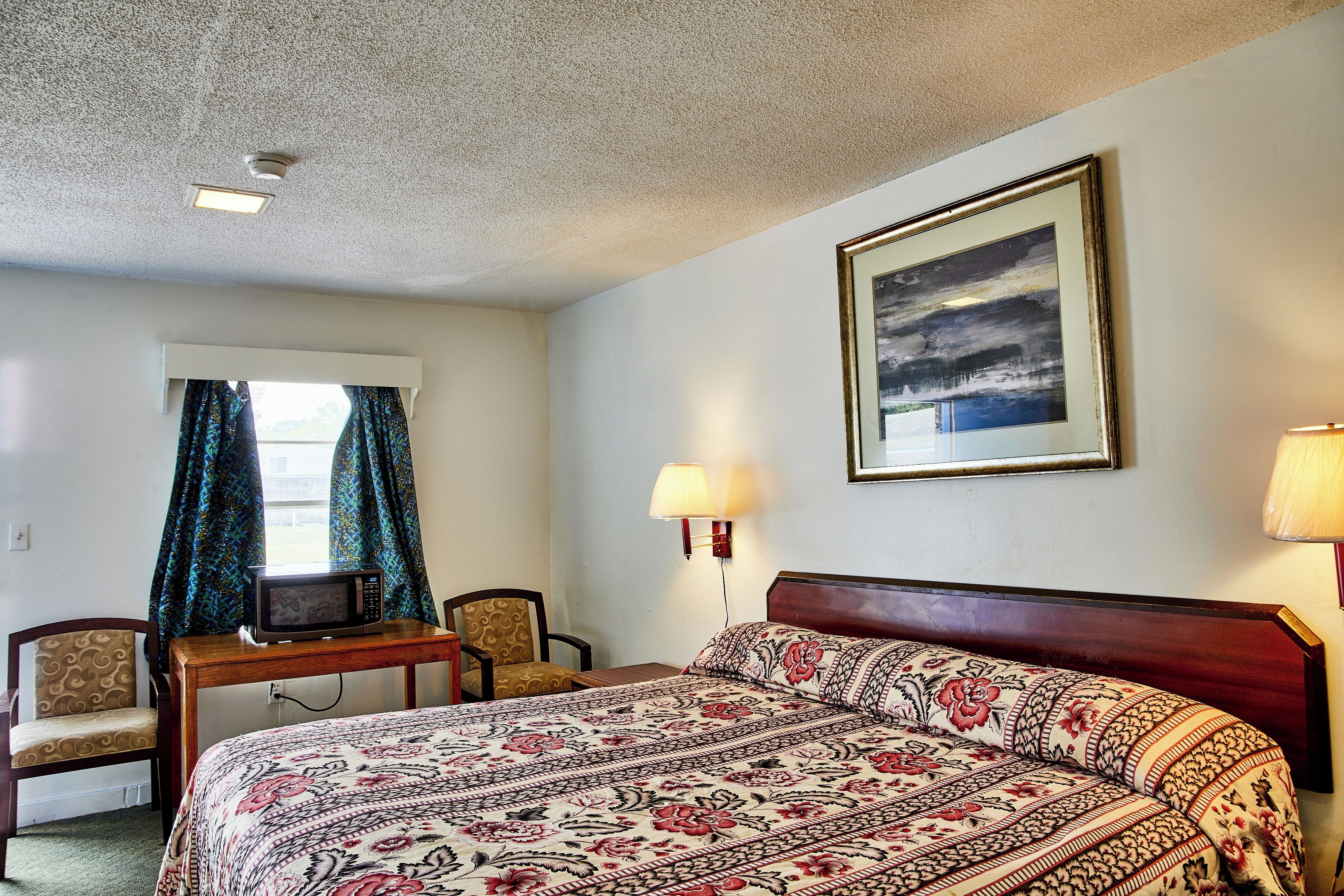 A hotel room with a bed, a desk with a microwave, two chairs, a window with curtains, and a framed picture on the wall.