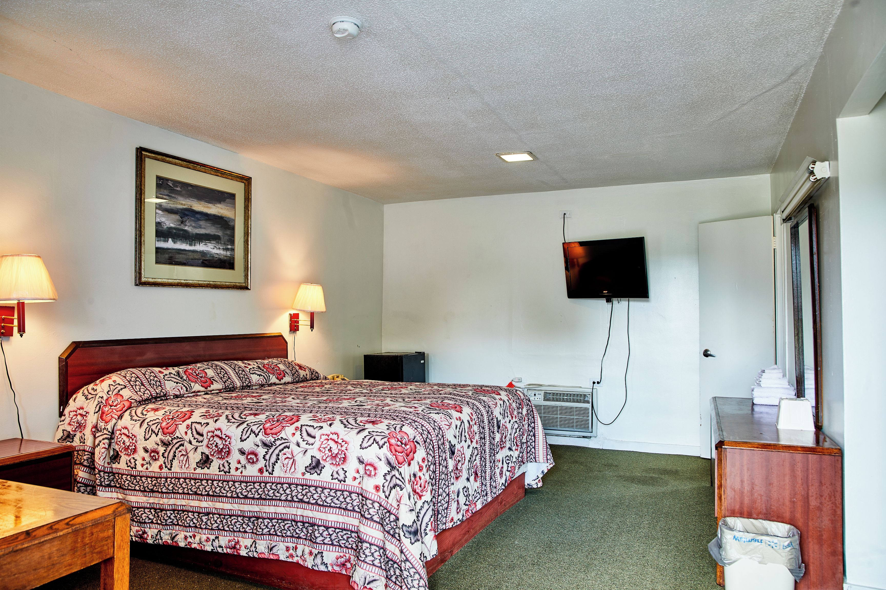 Quarto de hotel com uma cama grande, uma escrivaninha, uma cadeira, uma televisão na parede e um aparelho de ar condicionado.