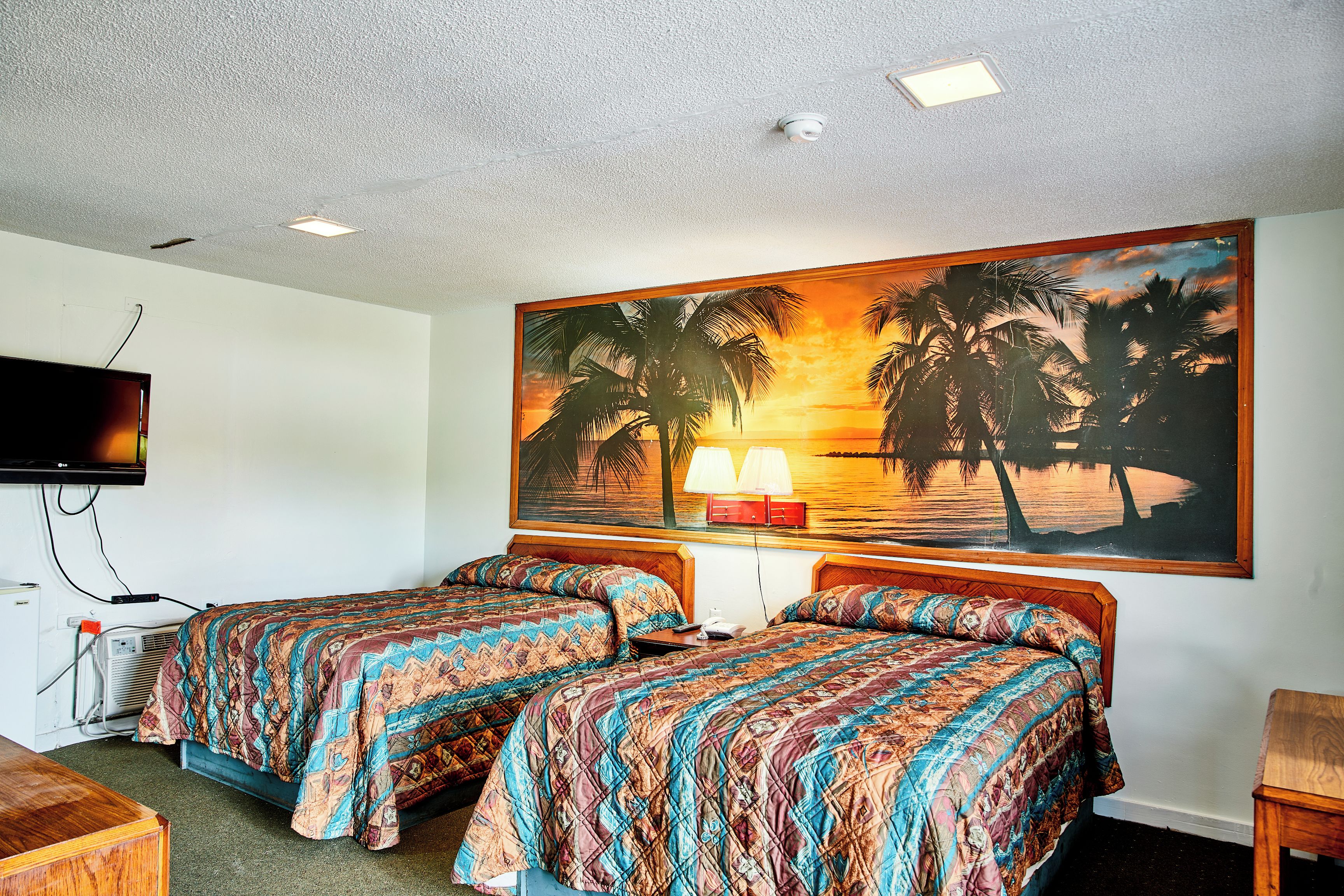 A hotel room with two beds, a TV, and a painting of palm trees at sunset.