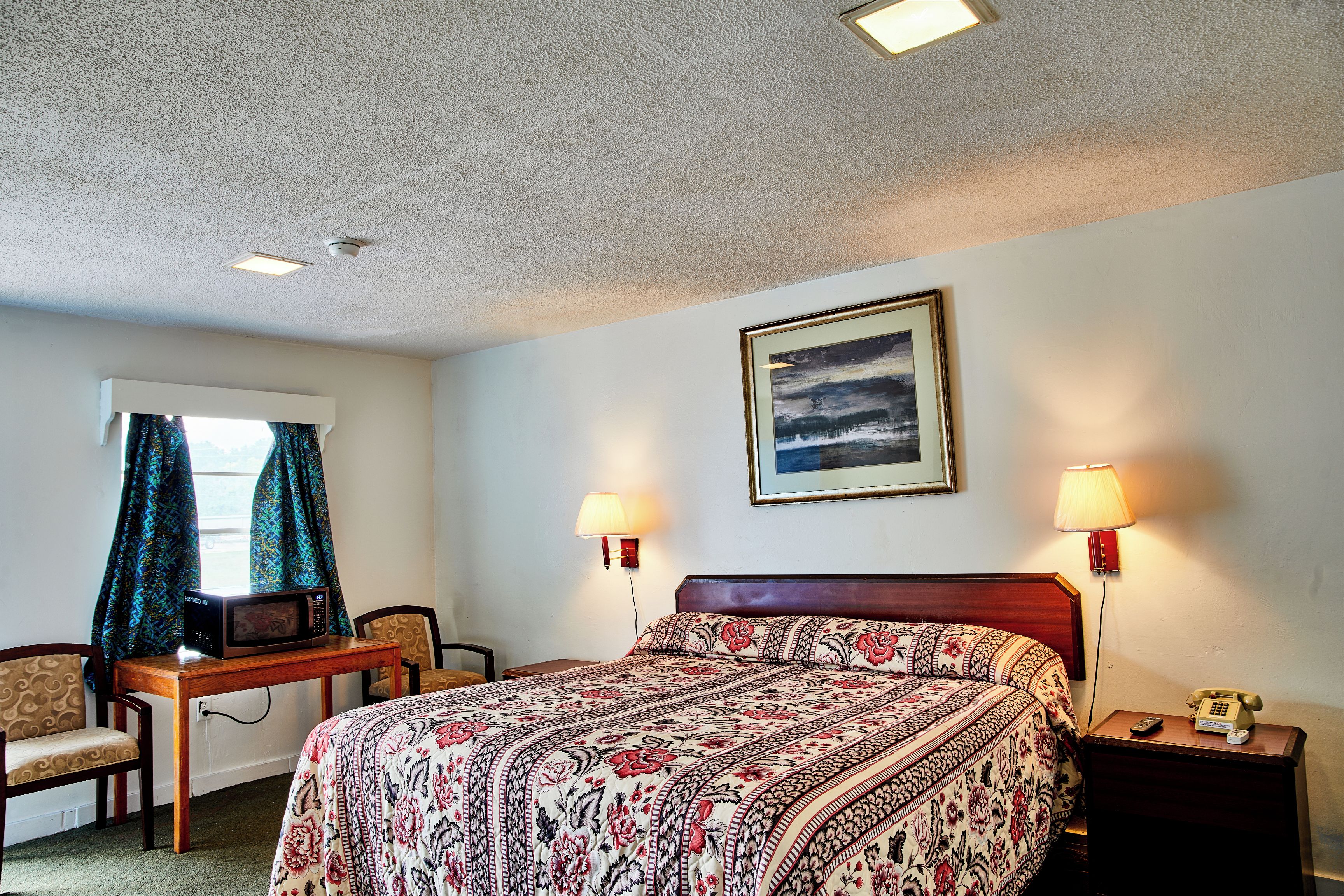 A hotel room with a bed, a desk with a microwave, two lamps, a chair, a nightstand with a phone, and a framed picture on the wall.