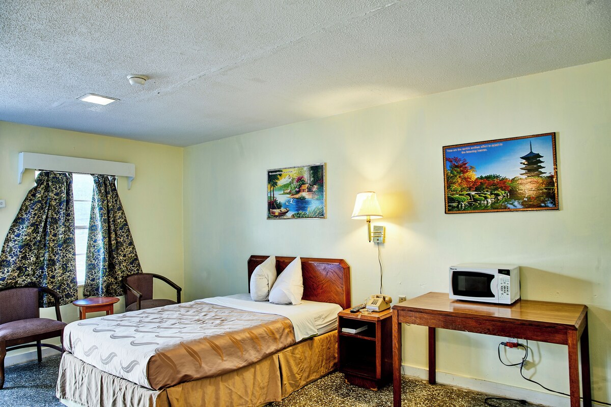 A hotel room with a bed, a desk, a chair, a lamp, and two paintings on the wall.