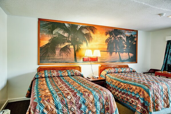 A hotel room with two beds, a painting of a sunset over palm trees, a nightstand with a lamp, and a desk with a chair. - Hospitality Inn by Hotel O Selma US 80 (Selma)