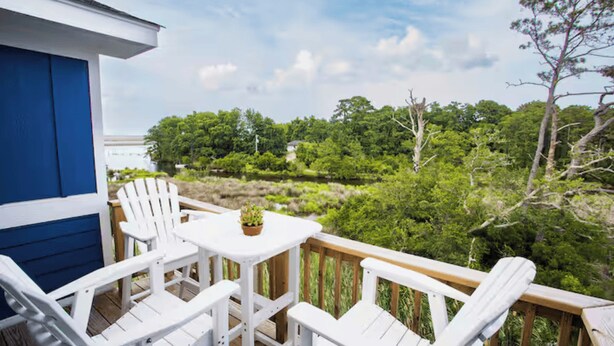 Una terraza de madera con sillas Adirondack blancas y una pequeña mesa, con vistas a un paisaje verde exuberante.