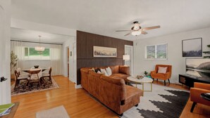 A modern living room with a brown sofa, a dining table with chairs, a ceiling fan, and a large window with blinds.