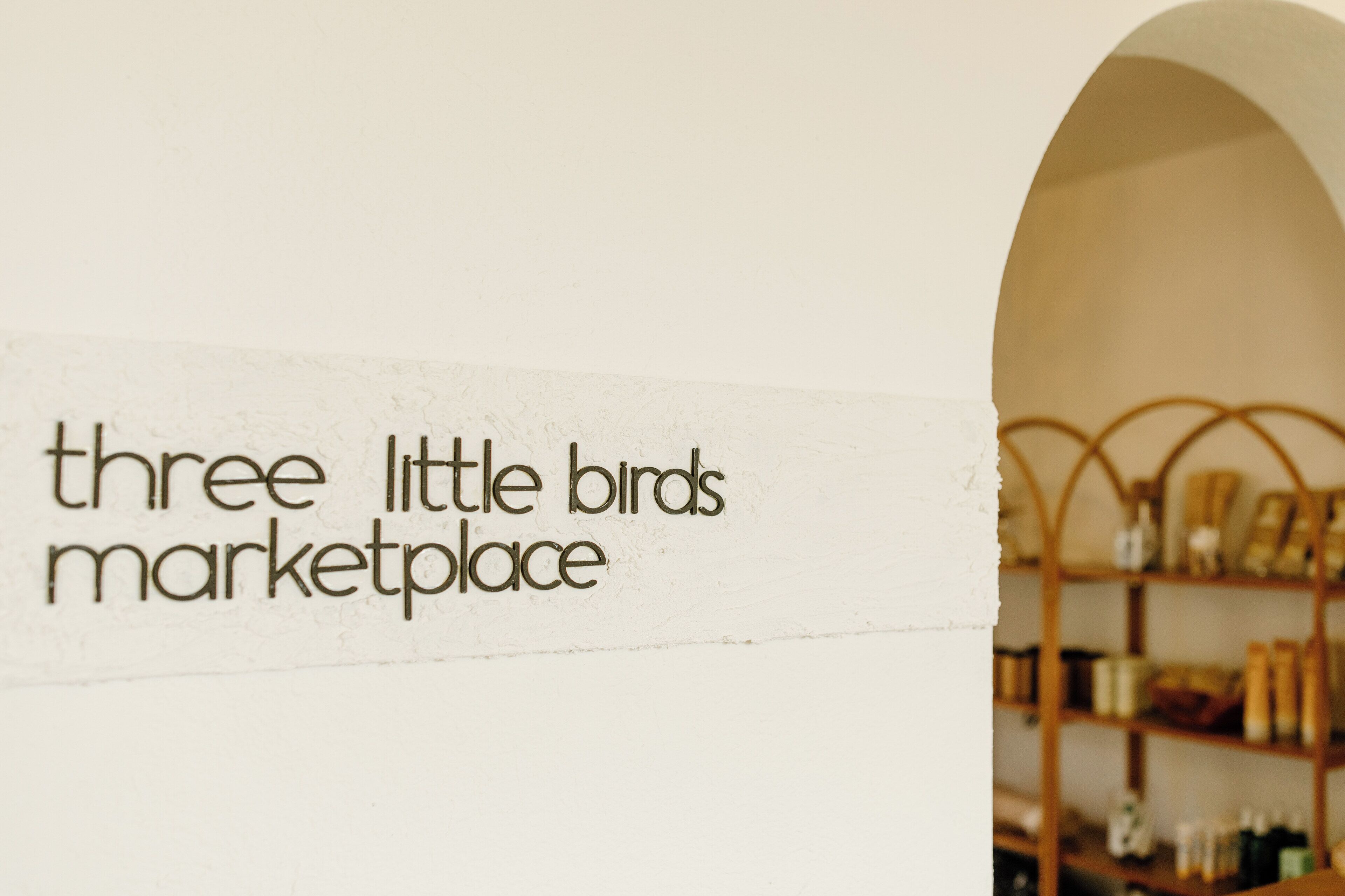 A sign reading "three little birds marketplace" with a wooden shelf in the background.