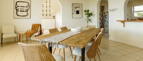 A spacious dining area with a long wooden table, wicker chairs, and a white wall with framed artwork.