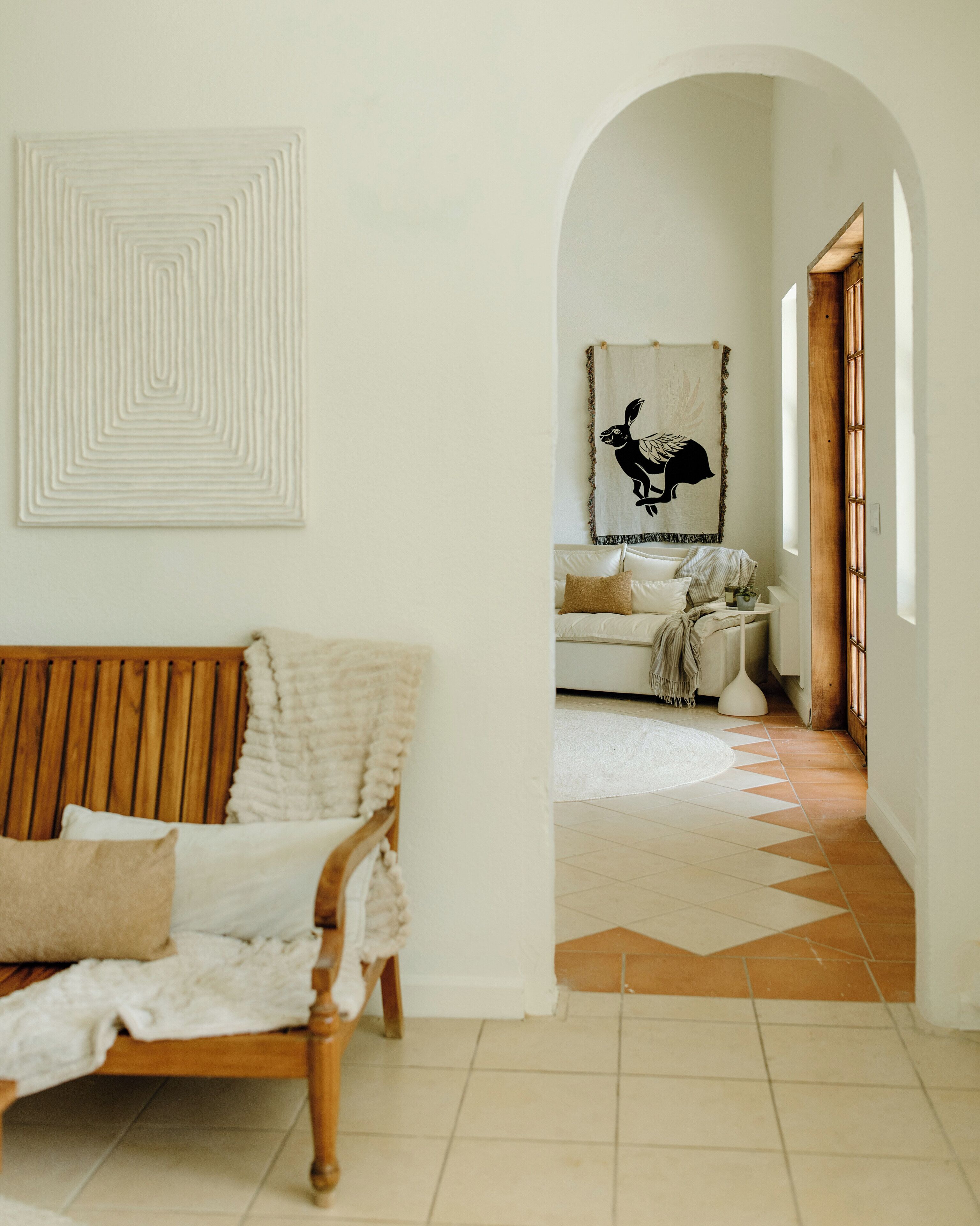 Une pièce avec un banc en bois, un canapé blanc, un tapis à motifs et un mur orné d’un motif géométrique.