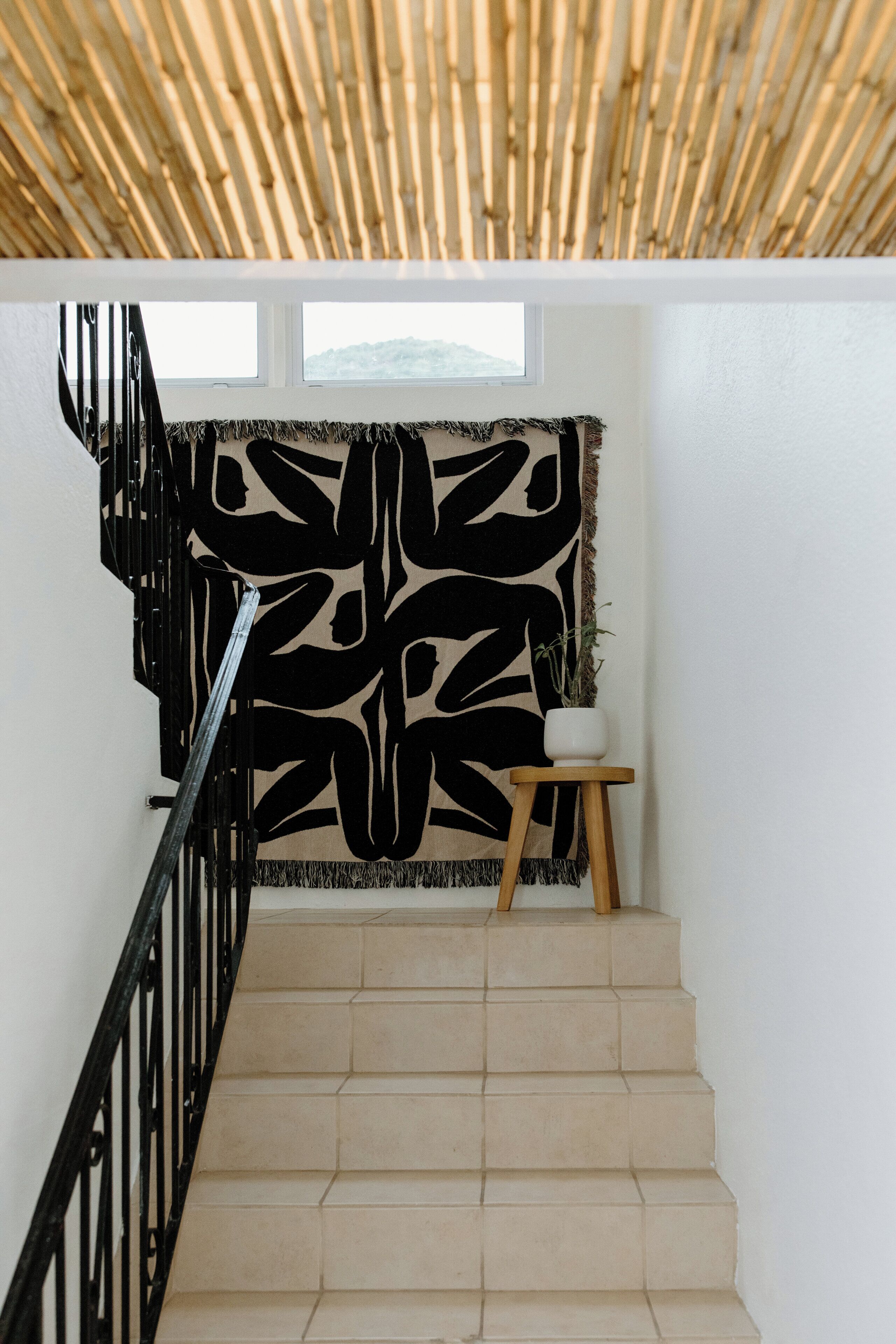 A staircase with a bamboo ceiling, a black abstract wall hanging, and a small wooden stool with a plant.