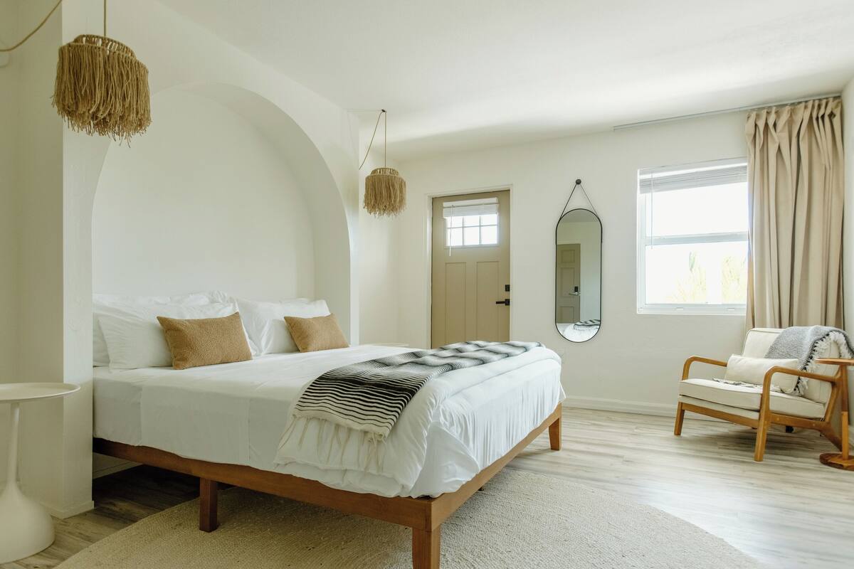 A bedroom with a large bed, a chair, a hanging light, and a mirror.