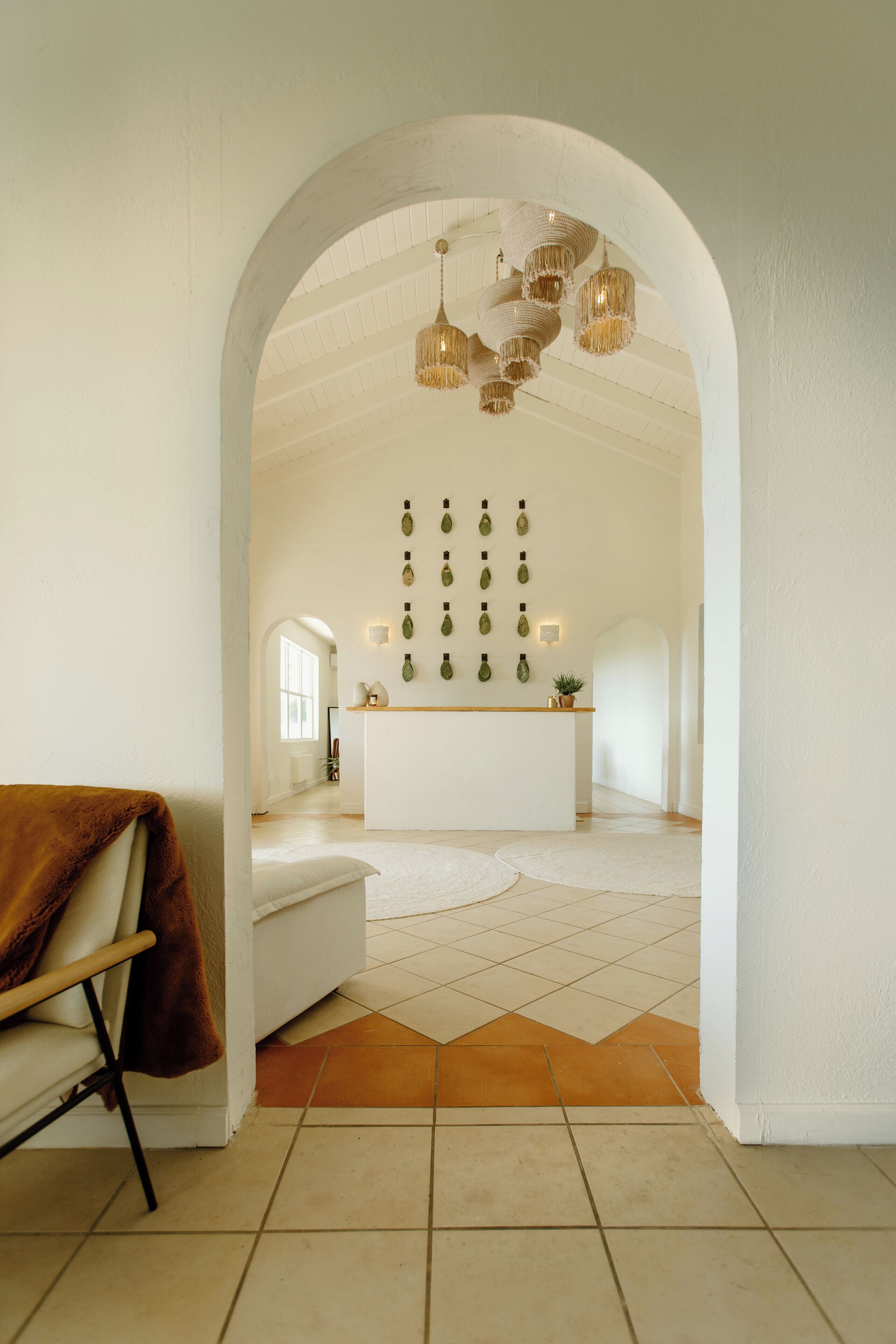 An interior with a white archway leading to a room with a white bench, a small table, and a wall with hanging lights.