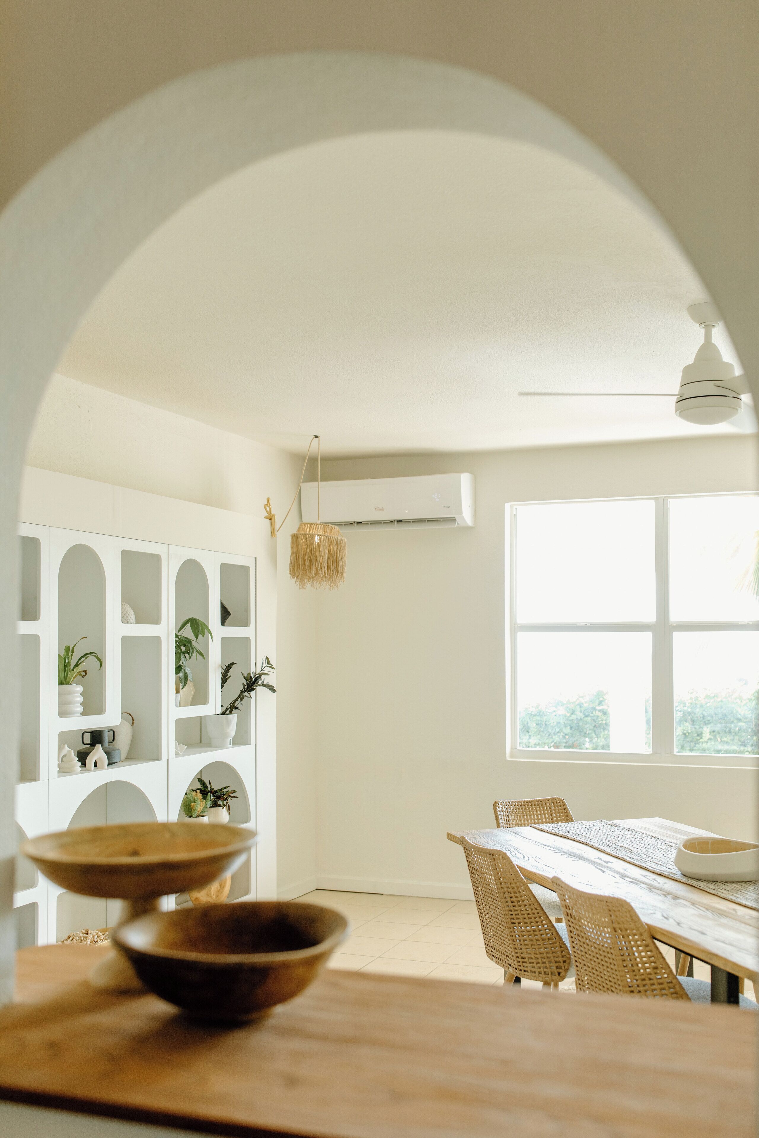 Un comedor con una mesa de madera, sillas de mimbre y un armario blanco con plantas.