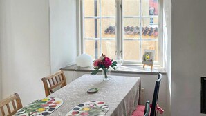 A dining area with a table set for two, a vase with flowers, and a window with a view of buildings. - Awesome home in Visby innerstad (Visby innerstad)